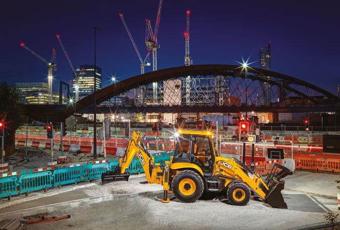 JCB 3CX Plus excavating with rear arm at night in front of a bridge